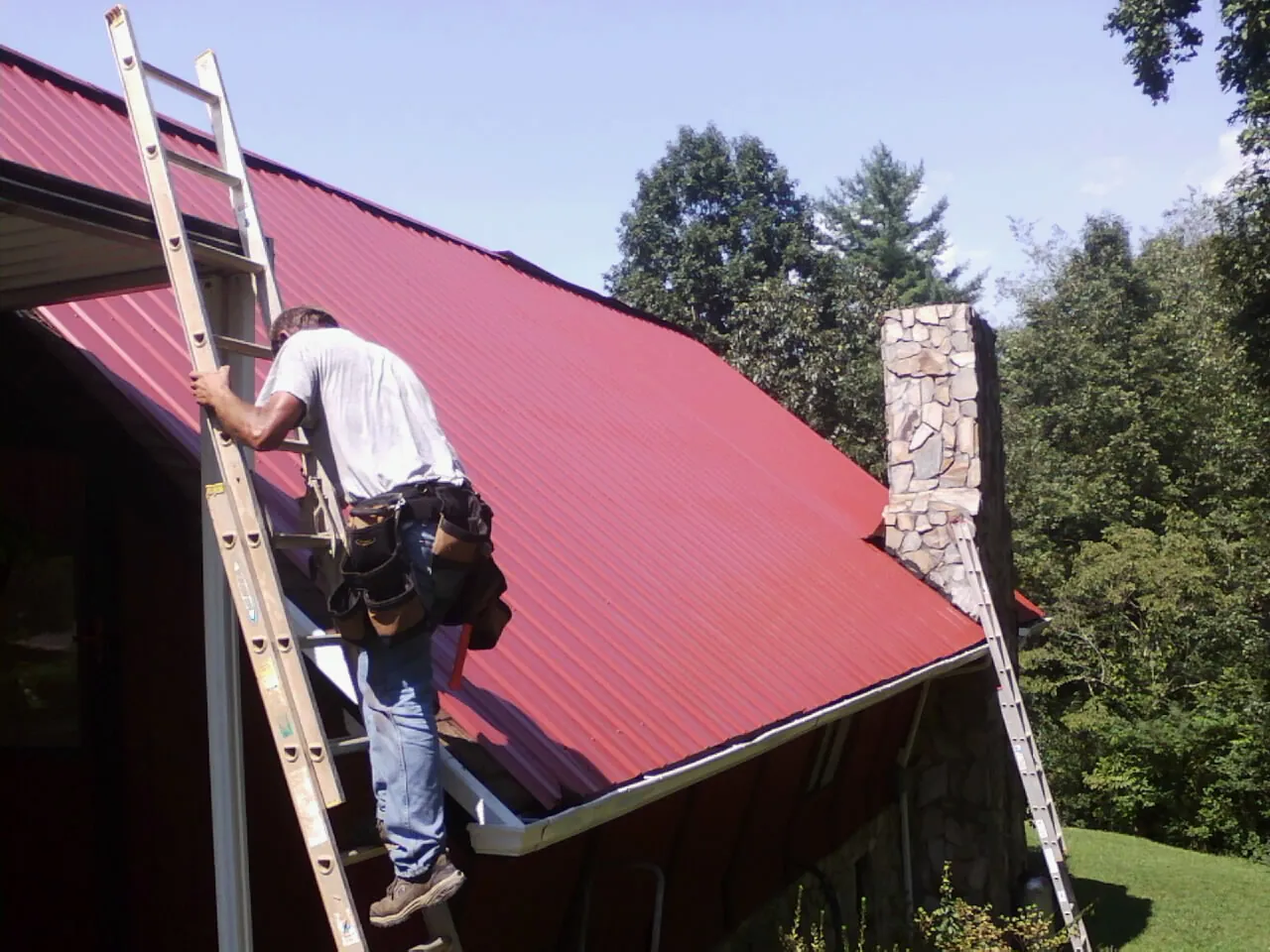 Roofer on a ladder beside a red metal roof and stone chimney — Soffit & Fascia Repair by Roofing Brothers in Park City